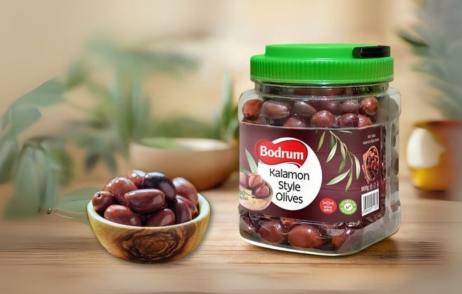 Jar of Bodrum Kalamon Style Olives on a wooden surface with olives beside it.